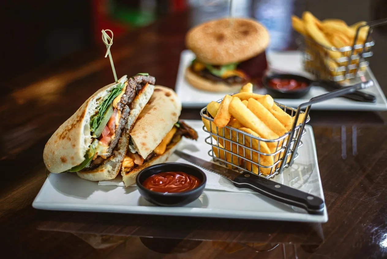 Loaded beef burger cut in half on a white plate with a wire basket of chips and tomato sauce, second burger in background